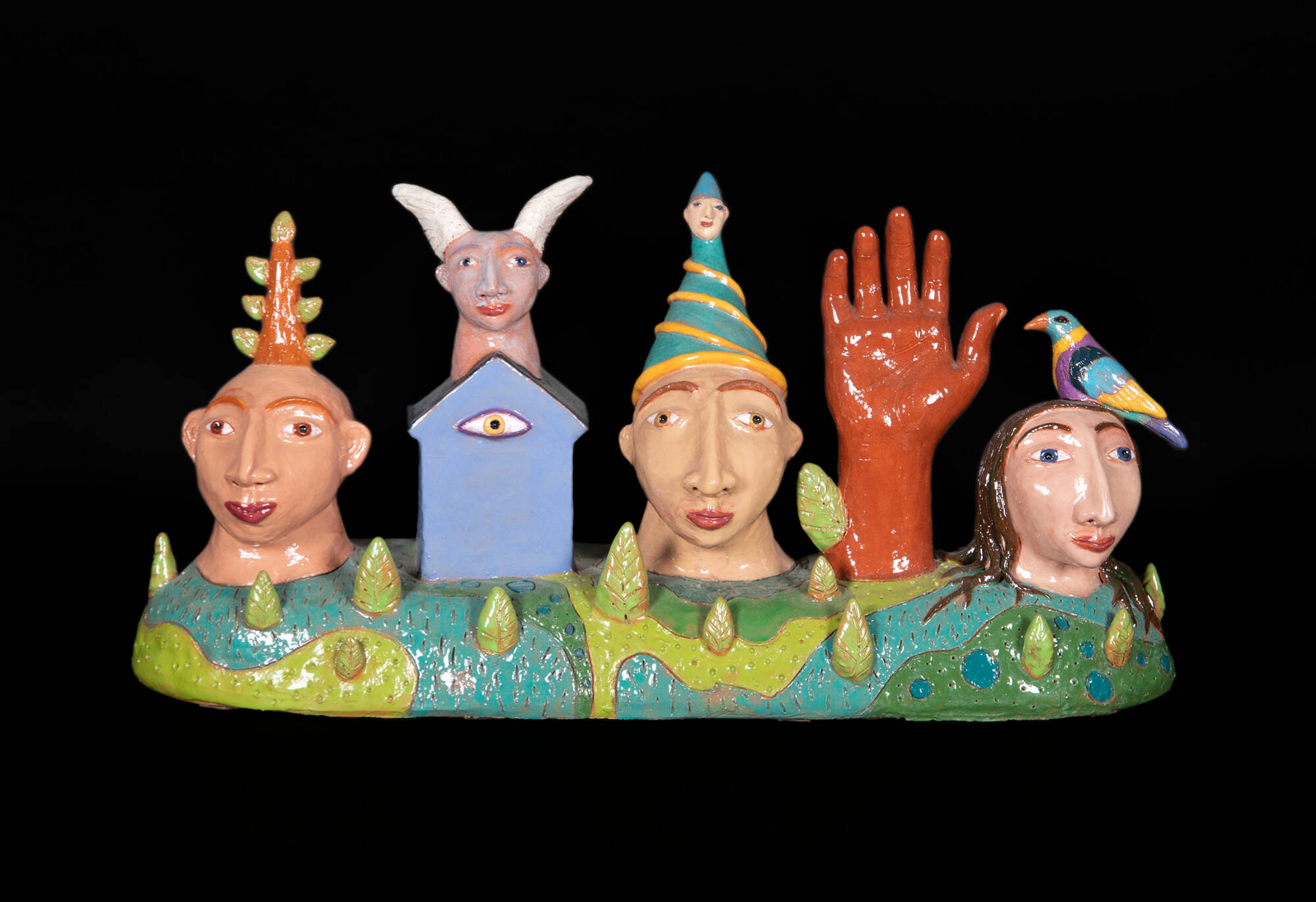 Colorful ceramic sculpture that has three heads, a house, and a hand situated on a base with elements of a landscape, like trees and blue water.